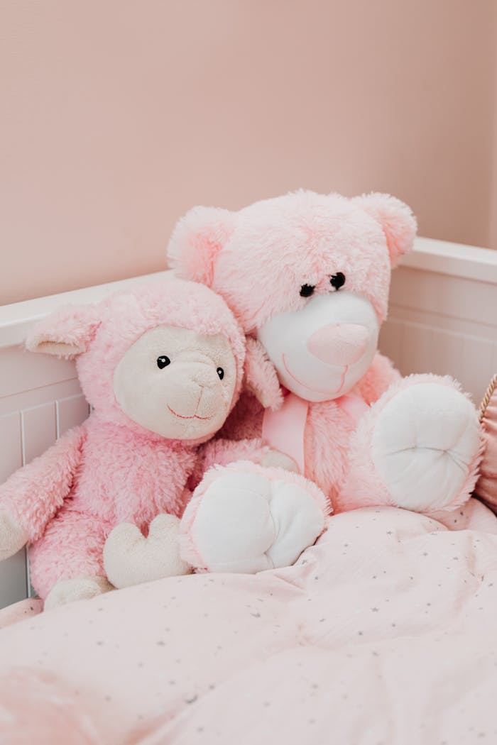 Two cute pink stuffed toys, a lamb and teddy bear, on a bed in a warm setting.